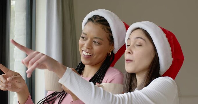 Seeing motion outside, Diverse women in Santa hats are eagerly pointing at view with mugs, laughing