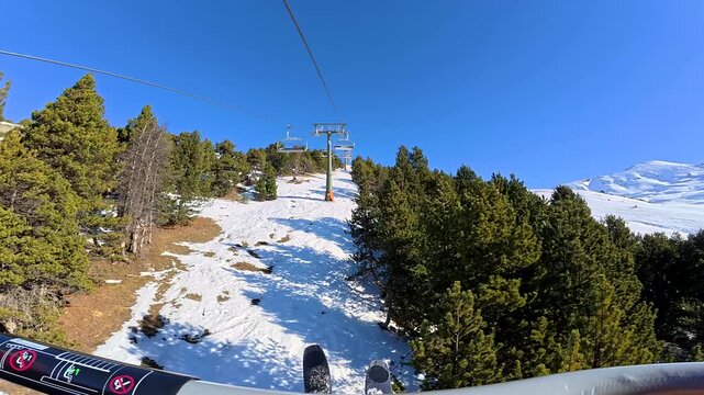 Point of view from a skier riding a chairlift up a sunlit, snowy mountain with pine trees, ascending towards the summit of a beautiful ski resort against a clear blue sky on a perfect winter day
