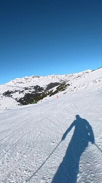 First person perspective of a skier descending a snowy slope at a ski resort, with the skier's shadow cast on the groomed piste under a clear blue sky and a beautiful mountain panorama