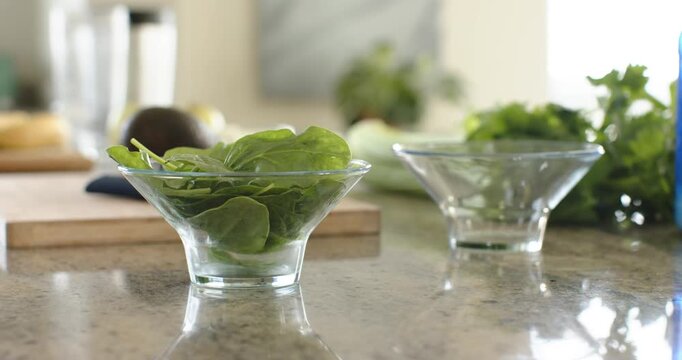 Camera is pushing in and adjusting focus, bringing glass bowl of spinach into focus on countertop