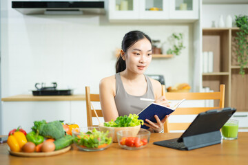 Asian woman in fitness wear uses tablet while eating healthy food at kitchen table. Holds tomatoes, writes in notebook. Clean eating and remote wellness lifestyle theme.