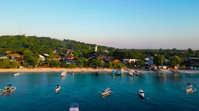 Drone video capturing a serene coastal town near Amed, Bali, featuring anchored boats and hilltop towers under clear blue skies. The panoramic view showcases vibrant colors and tranquil scenery.