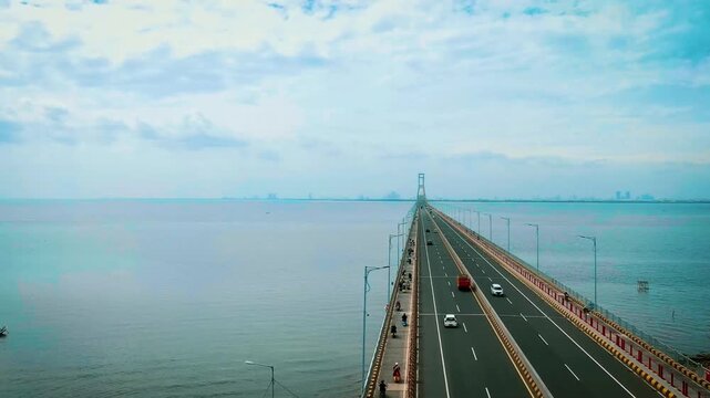 Aerial drone view of long bridge stretching over calm water with autumn colored trees and distant city skyline near Benoa Harbour, capturing wide coastal scene and smooth traffic flow in Bali