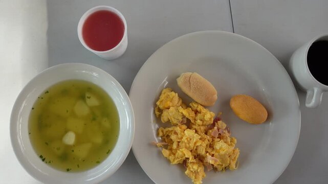 Bright overhead view of a typical Colombian breakfast featuring scrambled eggs, soup, bread, and beverages. Clean presentation with natural lighting creates a welcoming, inviting scene.