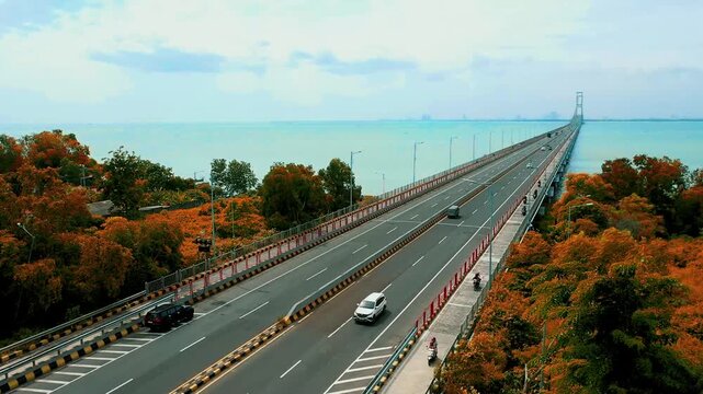 Aerial drone view of long bridge stretching over calm water with autumn colored trees and distant city skyline near Benoa Harbour, capturing wide coastal scene and smooth traffic flow in Bali