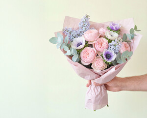 Hand holds pastel bouquet with roses, anemones, lilacs, and eucalyptus wrapped in pink paper. A delicate floral gift for wedding, anniversary, or seasonal celebration in spring or early summer.