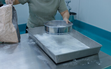 Unknown hand people sifting flour with sifter on stainless steel table in clean food preparation area, Factory production line baking preparation professional kitchen workflow industrial technology.