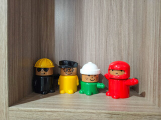 Four colorful vintage toy figures with simple features, dressed in different outfits and hats, are displayed on a wooden shelf