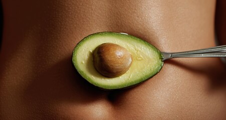 A halved avocado on a spoon is resting on a person's body, healthy eating.