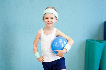 Confident young boy in a tank top and headband holds a blue exercise ball against a textured blue wall in a fitness studio.