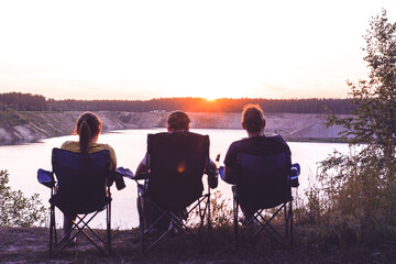 Three people silhouetted in chairs watch the peaceful sunset over a large lake from a high vantage point, enjoying the evening.