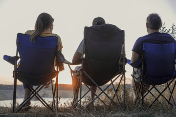Close-up, low-angle shot of three people silhouetted in chairs watching the sunset over a lake, emphasizing relaxation and togetherness.