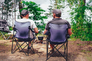 Two men relaxing in camping chairs, viewed from behind, watching a campfire with a cauldron at a serene forest campsite.
