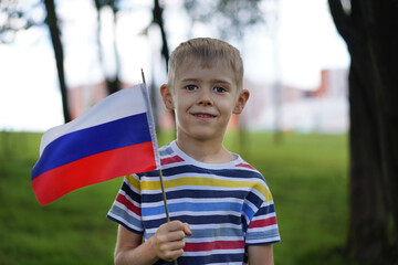 Smiling young boy holding a Russian flag in a striped shirt, standing in a sunny park with trees and blurred buildings behind him.