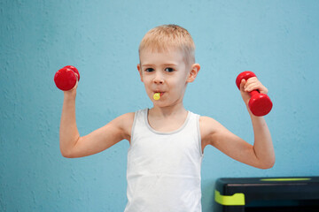  Young boy in a white tank top humorously poses, flexing with two red dumbbells while holding a candy, against a blue wall.
