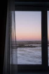Winter dawn landscape seen through a window frame with a sheer curtain, creating a serene and introspective view of a snow-covered field and distant forest at sunrise
