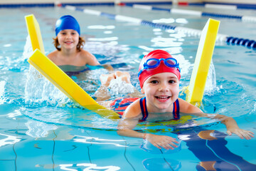 Portrait of Caucasian child girl wearing swim cap and goggles smiling at camera while swimming with pool noodle, another Caucasian child girl swimming in background in indoor pool
