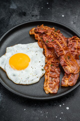 Crispy bacon and bright fried egg on black plate over dark textured background, perfect for breakfast, keto meals and gourmet food themes.