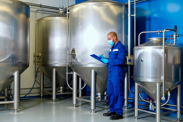 Man wearing protective mask and gloves inspecting stainless steel tanks in industrial facility, holding clipboard and writing notes, standing near large fermentation equipment