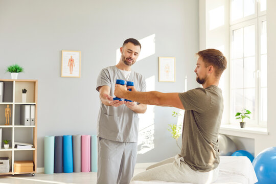 Young man doing sport exercise using dumbbells with support from male friendly nurse in clinic. Physiotherapist helping patient in lifting dumbbell. Physiotherapy and physical rehabilitation concept. - Powered by Adobe