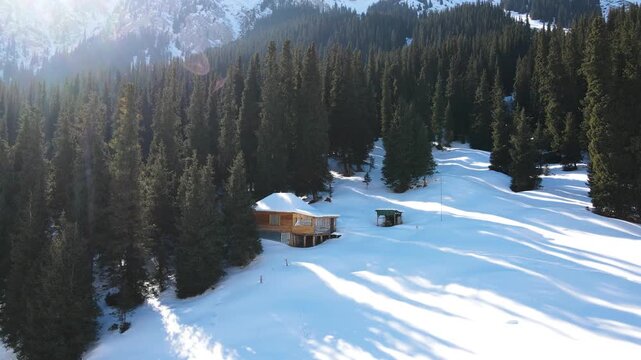 Aerial view of a secluded wooden cabin surrounded by pine trees in a snowy mountain landscape with a strong red sun flare