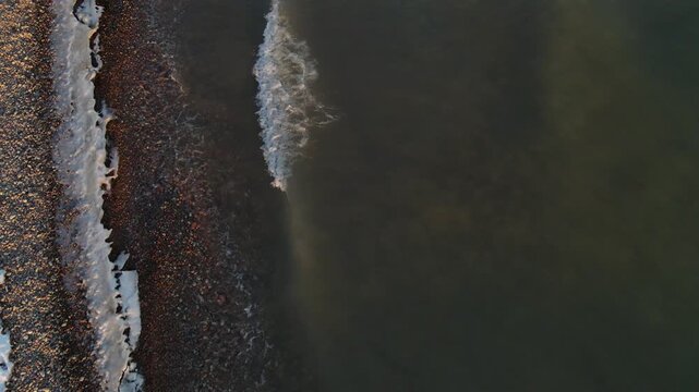 Aerial view of a deserted sandy beach covered in snow with rows of white sunshades and a long pier stretching into the blue water of Lake Issyk-Kul