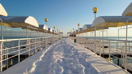 Perspective view of a long snow-covered pier with white canopies and yellow street lamps stretching into the blue water of Lake Issyk-Kul - Powered by Adobe