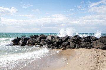Waves crash against line of dark, rugged rocks along sandy shoreline, turquoise water bright, cloudy sky, dynamic coastal scene.captures energy and beauty of the seaside, nature travel ocean landscape