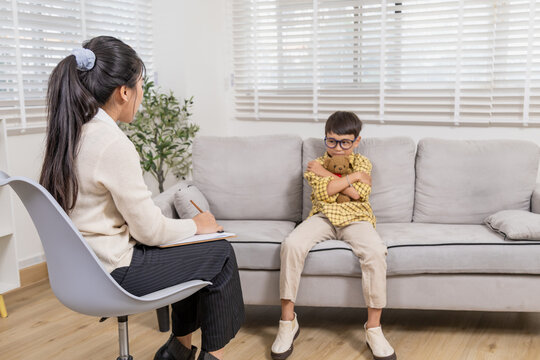 Young child sitting on sofa hugging teddy bear tightly, avoiding eye contact with therapist during counseling session, showing closed posture and emotional withdrawal in private consultation room