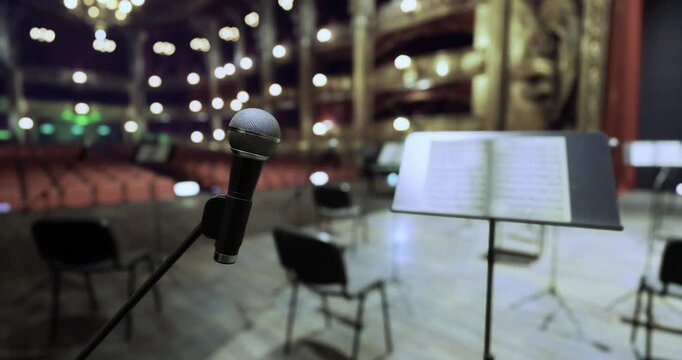 A microphone stands at the forefront in an ornate theater, surrounded by empty chairs and music stands, signaling a moment of anticipation before an evening concert.