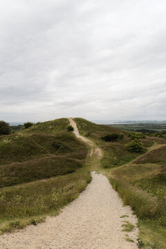 Winding gravel path leads over gentle, grassy hills, an overcast sky. Soft, muted light, natural landscape, calm and inviting scene ideal for themes of travel, exploration, nature, outdoor adventure