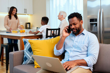 Black man using laptop and talking on smartphone sitting on sofa while Black woman, Black boy, and Caucasian senior woman interacting in kitchen background