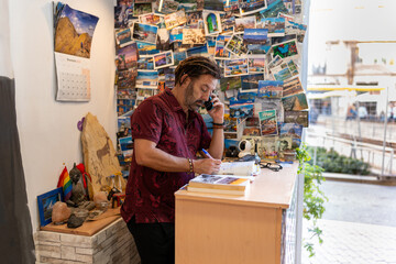 Male travel agent working in his office, talking on phone and writing in a notebook