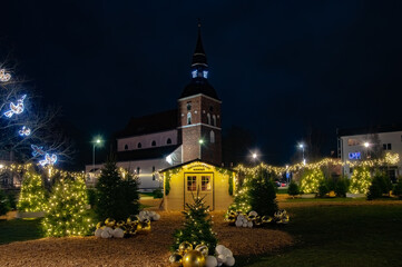 Valmiera,Latvia-December 04.2025-Festive Christmas lights, decorated trees, and a small holiday house glow warmly in front of a historic church on a winter night.