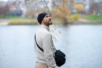Thoughtful man in a beanie and sweater looking up by a serene river on a cool autumn day. Ideal for themes of contemplation, nature, travel, and tranquility. © dizfoto1973