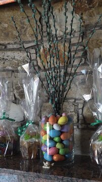 Colorful Easter eggs in glass vase on granite mantel, wrapped chocolate gifts in cellophane with green ribbon, tall twig branches arranged as rustic spring