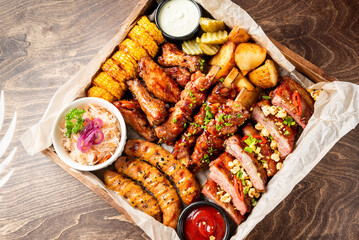 A delicious rustic assorted grilled meat and vegetable BBQ platter with various dipping sauces in a wooden serving tray on a dark wooden table