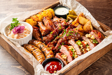 A delicious rustic assorted grilled meat and vegetable BBQ platter with various dipping sauces in a wooden serving tray on a dark wooden table