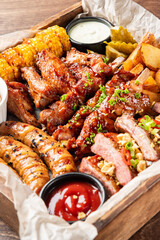 A delicious rustic assorted grilled meat and vegetable BBQ platter with various dipping sauces in a wooden serving tray on a dark wooden table