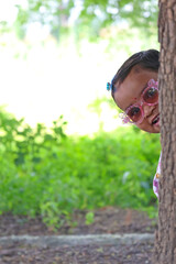 An Indian girl child wearing pink sunglasses peeking from behind a tree with a playful happy expression, standing outdoors in bright natural greenery with soft daylight creating a cheerful background