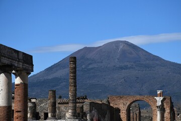 Mt Vesuvius rises up above the ruins of the Forum in Pompeii. A road can be seen zig-zagging across the mountain. The sky is blue.