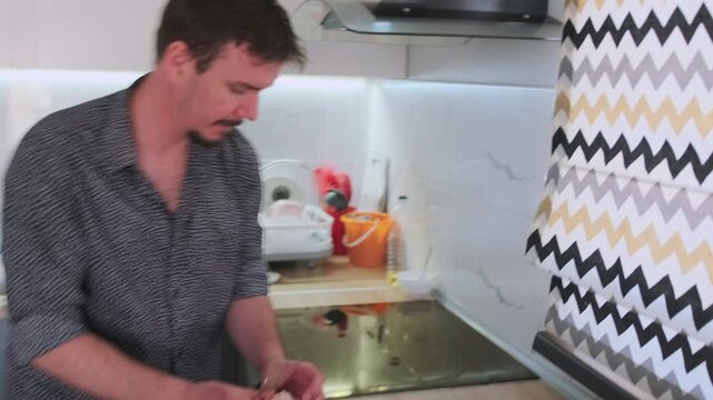 A man in a patterned shirt cracks an egg into a bowl, playfully dancing to music in a modern kitchen. Afterward, he throws the eggshell into the trash, blending cooking with fun and everyday routine.