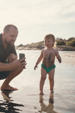 Father photographing son through smart phone at beach