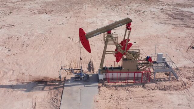Aerial drone captures oil pumpjack from directly above, showing industrial equipment in operation surrounded by barren desert land. Geometric forms, shadows, and sandy textures of the extraction site.