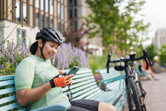 Smiling female cyclist using smart phone while sitting on bench at park