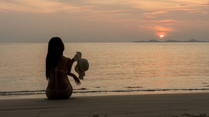 A woman sitting on the beach holding a hat while watching the warm sunset over the sea.