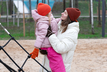 Happy young mother and adorable daughter hugging together at a playground, playing on a cold day dressed warmly clothes, tender moment, loving young mum and cute girl child cuddling, good relationship