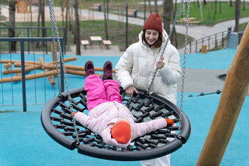Happy mother and daughter enjoying playful time together at a playground, with the daughter lying on a round swing while the mommy gently pushes. Full length carefree two generations family having fun