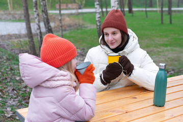Affectionate mother and daughter enjoying warm drinks together at a picnic table in the park, bonding outdoors on a cold day dressed warmly clothes, during family time tender moment. Pleasure be mommy