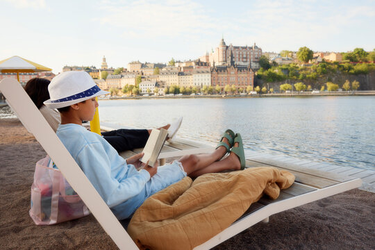 Full length of boy reading book while sitting comfortably on deck chair near river